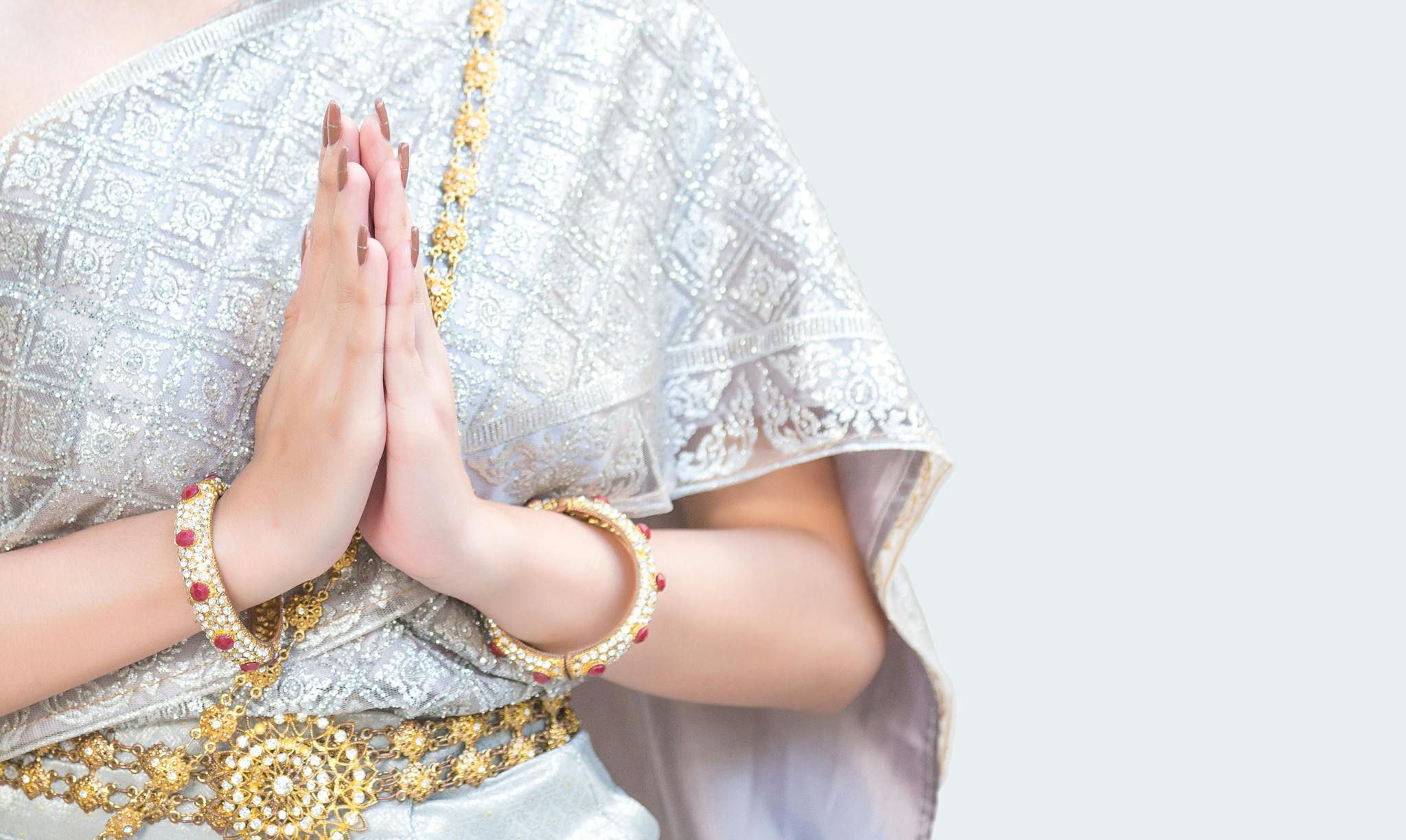 Close-up of a woman in traditional silver attire and gold jewelry folding her hands in a polite gesture.